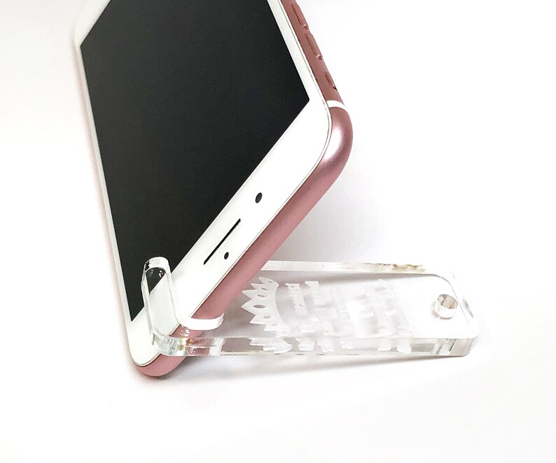 May include: A rose gold smartphone rests on a clear acrylic phone stand. The stand is designed to hold the phone upright. The phone's screen is black, and the stand has decorative cutouts.
