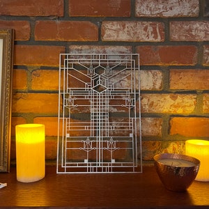 Pode incluir: Uma pe&ccedil;a de arte de parede de metal prateado com um design geom&eacute;trico, que se assemelha a um painel de janela. A arte &eacute; exibida em uma parede de tijolo com duas velas acesas e uma tigela de cobre sobre uma superf&iacute;cie de madeira.