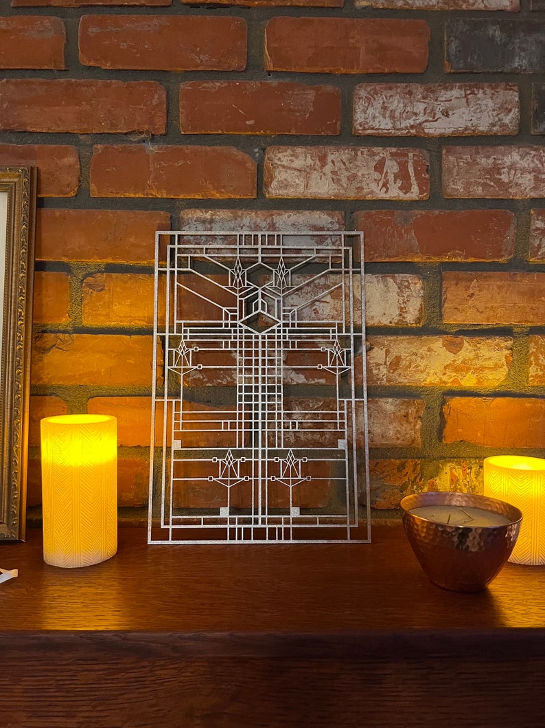 Pode incluir: Uma pe&ccedil;a de arte de parede de metal prateado com um design geom&eacute;trico, que se assemelha a um painel de janela. A arte &eacute; exibida em uma parede de tijolo com duas velas acesas e uma tigela de cobre sobre uma superf&iacute;cie de madeira.