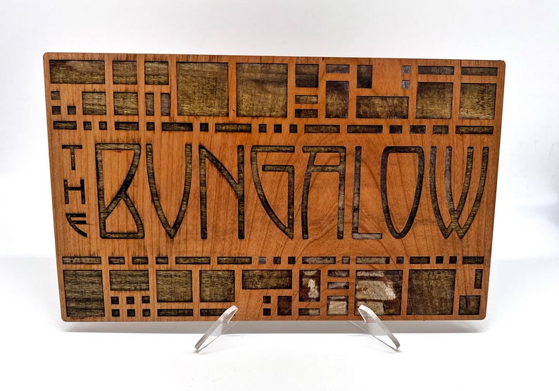 May include: Wooden sign with the text "THE BUNGALOW" carved into the wood. The sign is rectangular with a light brown finish and a geometric pattern around the text. The sign is displayed on a clear stand.