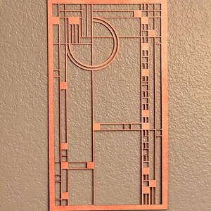 May include: A copper-colored, rectangular wall art piece with a geometric design. The artwork features a series of straight lines, squares, and a circle, creating an abstract pattern. The piece is mounted on a textured, neutral-toned wall.