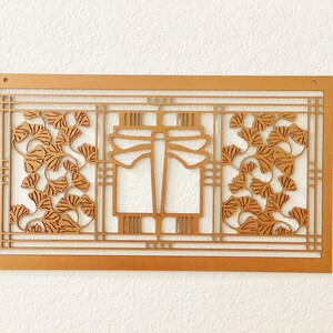 May include: A wooden wall art piece with a geometric design. The artwork features a rectangular frame with a central dragonfly motif and stylized ginkgo leaves. The piece is made of light brown wood and is mounted on a white wall.