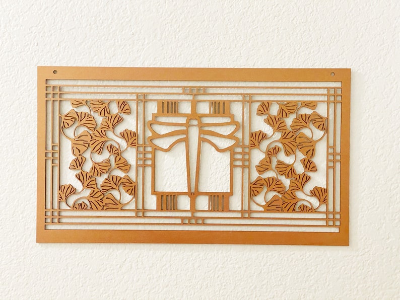 May include: A wooden wall art piece with a geometric design. The artwork features a rectangular frame with a central dragonfly motif and stylized ginkgo leaves. The piece is made of light brown wood and is mounted on a white wall.