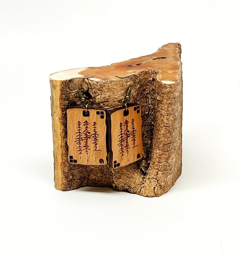 K&ouml;nnte beinhalten: Ein Paar Holzohrringe mit einem Waldmotiv. Die Ohrringe sind rechteckig und haben eine braune Holzoptik. Das Waldmotiv ist in das Holz ge&auml;tzt und zeigt eine Vielzahl von B&auml;umen. Die Ohrringe h&auml;ngen an einem Holzstamm.