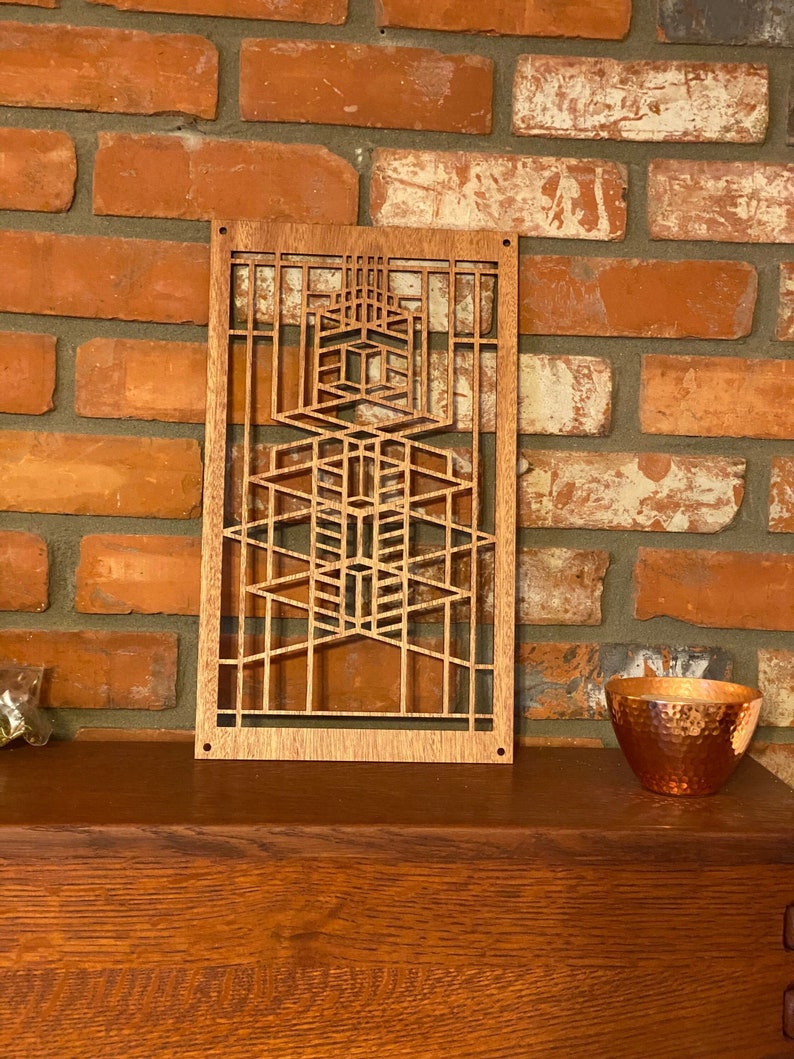 May include: A wooden wall art piece with a geometric design. The art piece is light brown and features interconnected rectangular and trapezoidal shapes. It is mounted on a brick wall. A copper bowl is on the right.