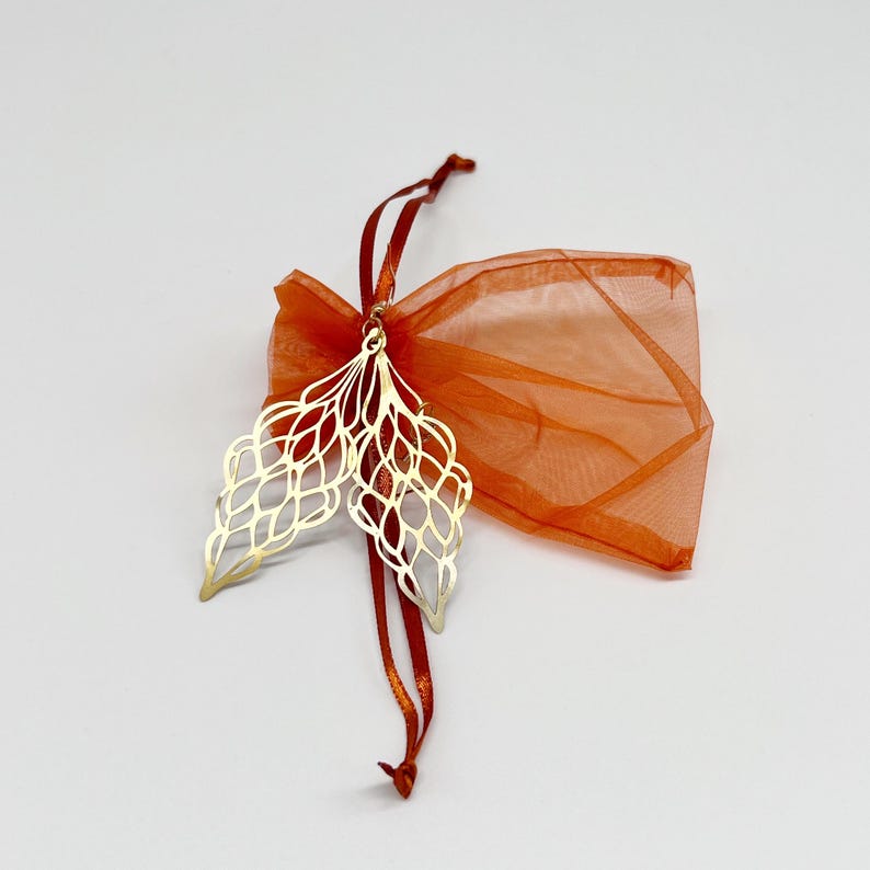 May include: A pair of gold metal bookmarks shaped like leaves with intricate cut-out designs. The bookmarks are packaged in a small orange organza bag with a red ribbon.