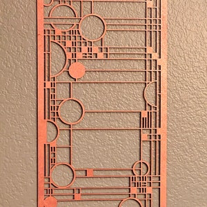May include: A copper-colored, rectangular wall art piece with a geometric design. The art features a series of circles, squares, and straight lines, creating an abstract pattern. The piece is mounted on a neutral-colored wall.