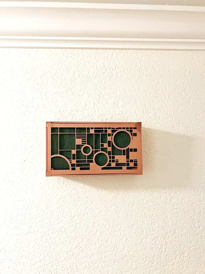 Op de afbeelding: Een rechthoekig koperkleurig wandkunstwerk met een geometrisch ontwerp van cirkels, vierkanten en lijnen. De binnenkant heeft een donkergroene stof. Het kunstwerk is bevestigd aan een getextureerde witte muur.