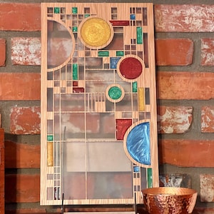 May include: A wooden wall art piece with a geometric design featuring colorful glass inserts. The design includes circles, squares, and rectangles in shades of blue, green, red, and yellow. The piece is displayed on a wooden shelf with a copper bowl.