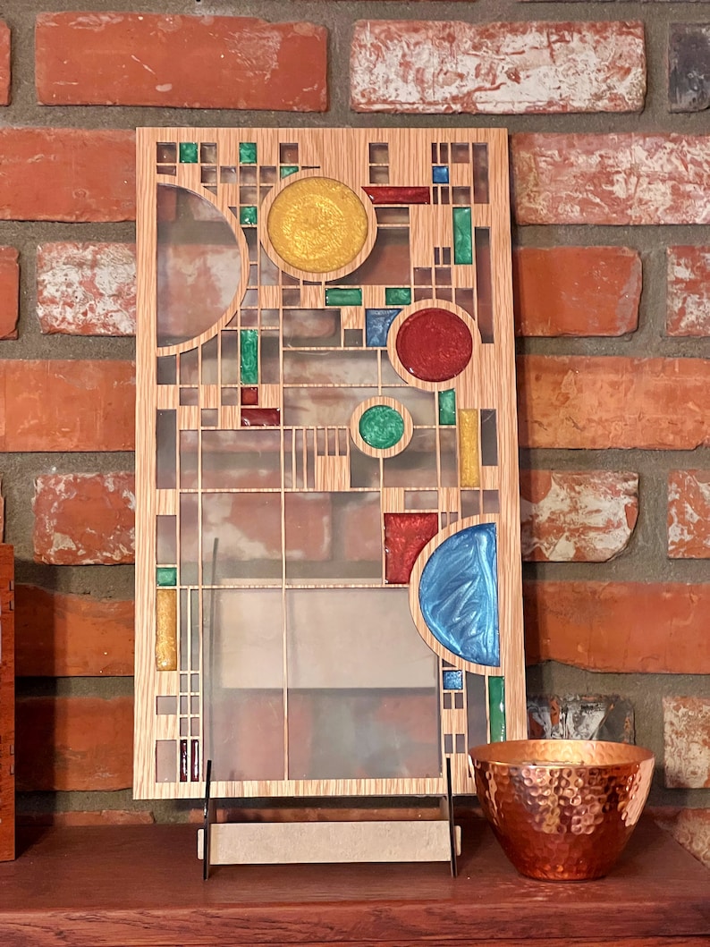 May include: A wooden wall art piece with a geometric design featuring colorful glass inserts. The design includes circles, squares, and rectangles in shades of blue, green, red, and yellow. The piece is displayed on a wooden shelf with a copper bowl.