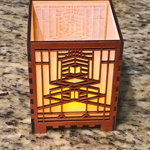 May include: A brown wooden candle holder with a geometric design. The candle holder has a square shape with intricate cutouts that allow light to shine through. The interior has a warm, yellow glow, suggesting a lit candle.