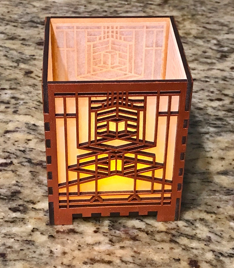 May include: A brown wooden candle holder with a geometric design. The candle holder has a square shape with intricate cutouts that allow light to shine through. The interior has a warm, yellow glow, suggesting a lit candle.