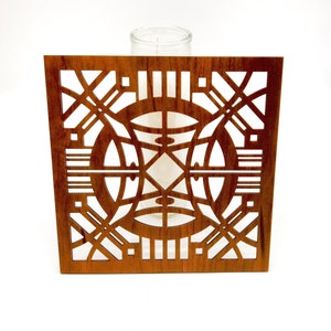 May include: A square wooden candle holder with a detailed geometric design. The holder is a warm brown colour and features cut-out patterns of circles, lines, and angles. A clear glass candle holder is visible behind the wooden frame.