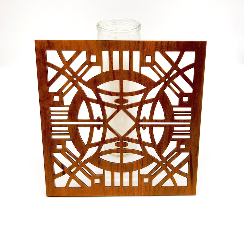 May include: A square wooden candle holder with a detailed geometric design. The holder is a warm brown colour and features cut-out patterns of circles, lines, and angles. A clear glass candle holder is visible behind the wooden frame.