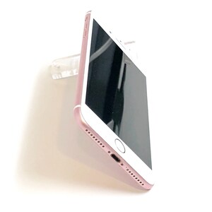 May include: Rose gold smartphone with a black screen, featuring a home button and speaker holes. The phone is mounted on a clear plastic holder, showcasing its sleek design against a white background.