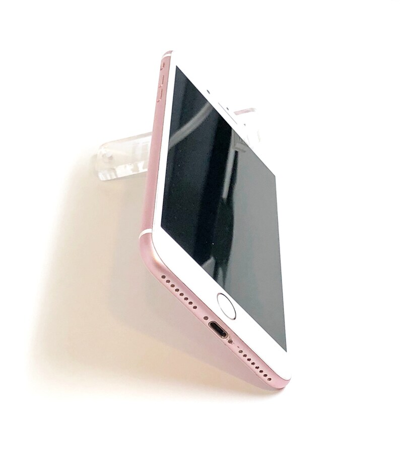 May include: Rose gold smartphone with a black screen, featuring a home button and speaker holes. The phone is mounted on a clear plastic holder, showcasing its sleek design against a white background.
