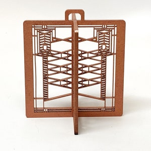 May include: A brown, laser-cut wooden decorative piece with an architectural design. The piece features a rectangular frame with intricate geometric patterns and a handle at the top. The design is symmetrical and stands upright.