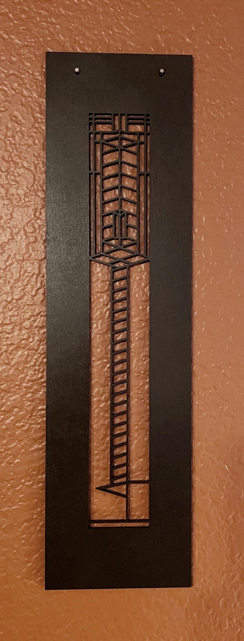May include: A dark brown, rectangular wall art piece with a geometric design. The art features a central vertical panel with a ladder-like pattern and a series of rectangular shapes. Two small silver rivets are visible at the top.