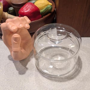 May include: A clear glass cloche with a round base and a domed lid. A lifelike, flesh-toned hand sculpture is positioned next to the cloche. A wooden bowl filled with colorful wax fruits is in the background.