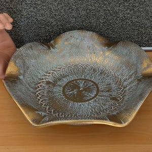 May include: A decorative ceramic ashtray with a scalloped edge and a white and gold design. The ashtray has a circular pattern in the center.