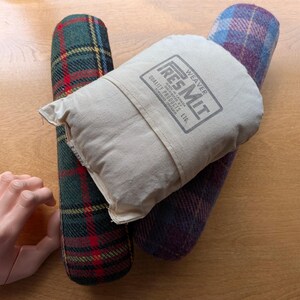 May include: Two cylindrical tailor's hams with plaid fabric patterns, one in green and red and the other in blue and purple, partially covered by a beige canvas pressing mitt with the printed text WEAVER RES MIT QUALITY PRODUCTS LTD.