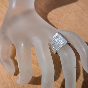 May include: A silver ring with a textured, geometric design. The ring is wide and has a square shape.