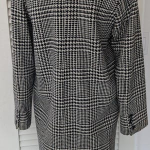 Puede incluir: Un abrigo largo con estampado de pata de gallo en blanco y negro. El abrigo tiene un cuello con solapas, pu&ntilde;os abotonados y una abertura en la espalda. La tela tiene una textura tejida cl&aacute;sica. Un abrigo de estilo cl&aacute;sico.