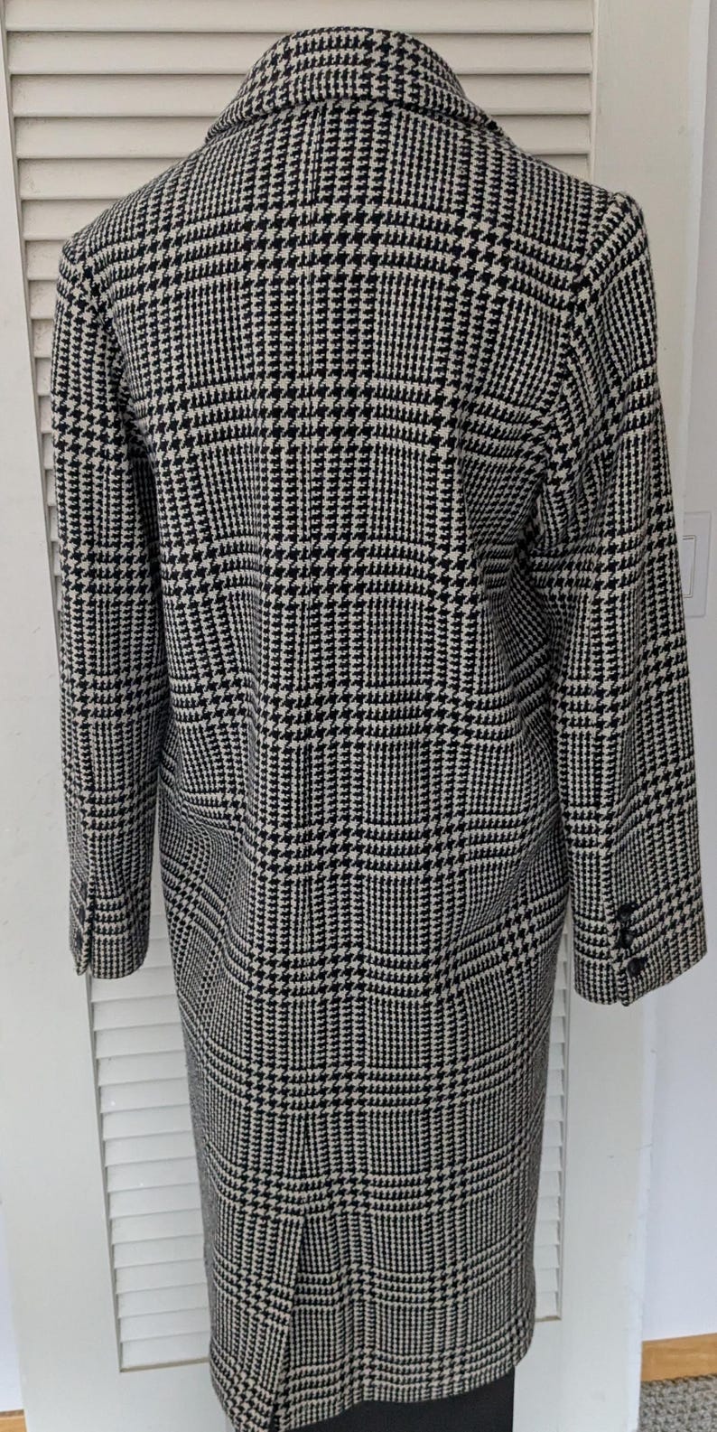 Puede incluir: Un abrigo largo con estampado de pata de gallo en blanco y negro. El abrigo tiene un cuello con solapas, pu&ntilde;os abotonados y una abertura en la espalda. La tela tiene una textura tejida cl&aacute;sica. Un abrigo de estilo cl&aacute;sico.