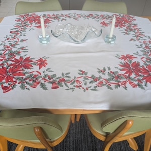 May include: A white tablecloth with a festive Christmas design featuring red poinsettias, green holly, and red ribbons. Two white candles in blue glass holders and a clear glass bowl are on the table. The table is surrounded by green chairs.