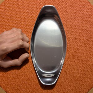 May include: A stainless steel oval serving dish with a polished finish. The dish has a shallow, elongated shape with rounded ends. The dish is on an orange woven placemat.
