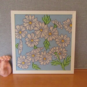 May include: A framed cross-stitch embroidery featuring a blue background with white flowers and green leaves.