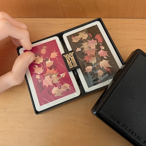 May include: Two decks of KEM plastic playing cards featuring a maple leaf design in a black storage case. One deck has a deep red background and the other has a black background, both with gold and orange leaves. The word MAPLE is printed on each.