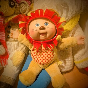May include: A vintage doll dressed as a lion, featuring a red and yellow mane, a smiling face, and a red polka-dot belly. The doll is part of a collection of plush toys, including a monkey and a Santa Claus figure.