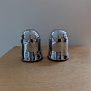 May include: Two shiny, silver salt and pepper shakers. The shakers have a bell-like shape with a flared base and a small hole at the top. They are reflective, mirroring the surrounding environment. The shakers are on a light brown wooden surface.