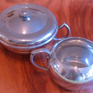 May include: A silver sugar bowl and creamer set with a lid on the sugar bowl. The set is on a wooden surface.