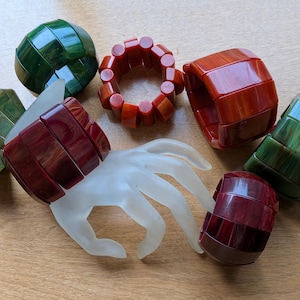 May include: A collection of six chunky bracelets in various shades of green, red, and brown, featuring smooth, segmented, and rounded designs. One bracelet is displayed on a translucent white hand-shaped stand, while the others are arranged around it.