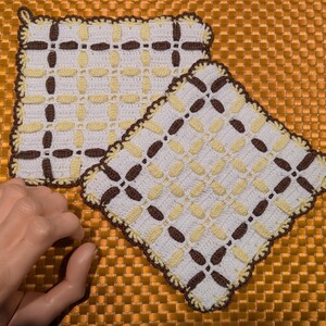 May include: Two handmade crochet potholders. The potholders are square with a white base, featuring a repeating pattern of brown and yellow stitches. The edges are scalloped and trimmed in brown. One potholder has a loop for hanging.