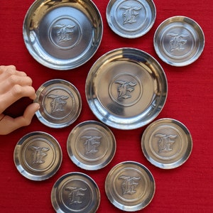 May include: Nine silver-colored coasters with a raised letter 'F' in the center. The coasters are arranged in a circle on a red surface.