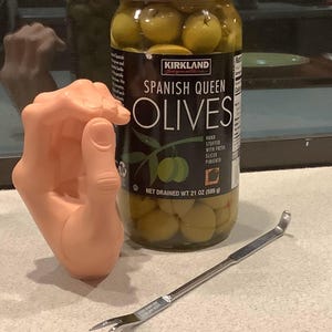 May include: A glass jar of green Spanish Queen olives with a label that reads "Kirkland Signature Spanish Queen Olives". The jar is sitting on a white surface next to a silver olive pitter.