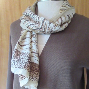 May include: A white and brown patterned scarf with a geometric design. The scarf is draped around a brown sweater.