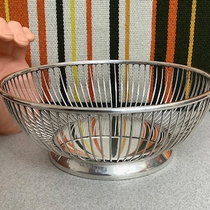 May include: A silver-toned metal wire bowl featuring a rounded, open-frame design with vertical slats and a solid circular base. The bowl has a polished finish and a wide, shallow shape, constructed from thin, evenly spaced metal rods.