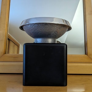 May include: A decorative bowl with a hammered texture and a silver rim, resting on a black square base. The bowl has a unique, textured surface and a metallic finish. The design is modern and artistic.