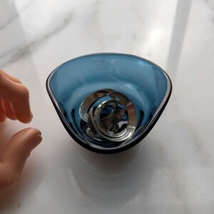 May include: A small, triangular blue glass bowl with a black rim. Inside the bowl are several silver-colored metal rings. The bowl is sitting on a white surface.