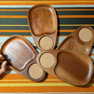 May include: Three wooden snack trays with a smooth, polished finish. Each tray features a large, rounded, recessed section for food and a smaller, circular cutout containing a light brown, textured cork coaster insert for a beverage.