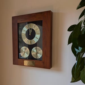 May include: A rectangular wall-mounted wooden clock featuring a dark brown frame, a central gold-toned clock face, and two smaller circular gold-toned gauges below. A small brass plaque at the bottom displays engraved text in a serif font.