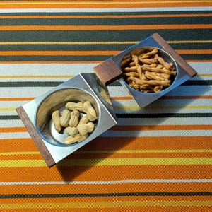 May include: Two square stainless steel bowls with dark wood accents on opposite corners. One bowl contains light brown peanuts in their shells, while the other bowl holds several thin, twisted, golden-brown baked snack sticks.