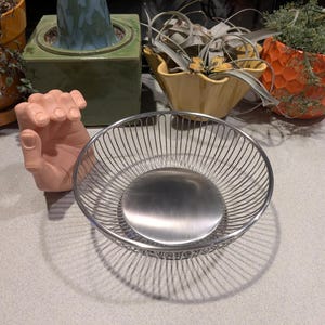 May include: A circular wire basket featuring a polished stainless steel base and thin, vertical metal rods forming the sides. The basket has an open-top design with a consistent, uniform spacing between each wire, creating a clean, metallic aesthetic.