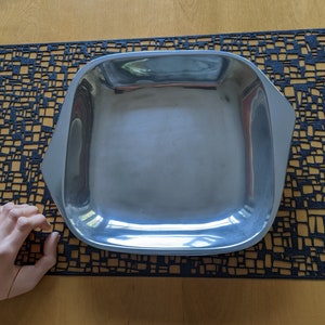 May include: A silver serving dish with a slightly rounded square shape. The dish has a smooth, reflective surface and a slightly raised rim.