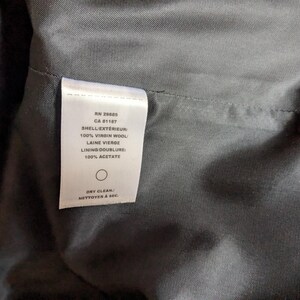 Puede incluir: Primer plano del interior de una prenda, que muestra una etiqueta blanca con texto. La etiqueta enumera detalles como "100% Lana Virgen" y "100% Acetato". La tela es de color gris oscuro y la etiqueta incluye instrucciones de limpieza en seco.