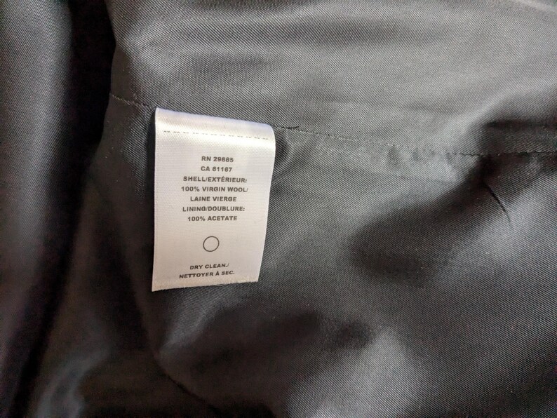 Puede incluir: Primer plano del interior de una prenda, que muestra una etiqueta blanca con texto. La etiqueta enumera detalles como "100% Lana Virgen" y "100% Acetato". La tela es de color gris oscuro y la etiqueta incluye instrucciones de limpieza en seco.